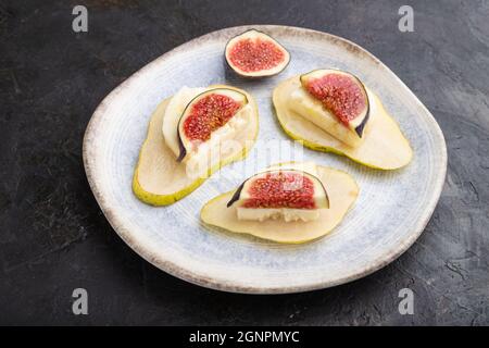 Sommervorspeise mit Birne, Quark, Feigen und Honig auf Keramikplatte auf schwarzem Betongrund. Seitenansicht, Nahaufnahme. Stockfoto