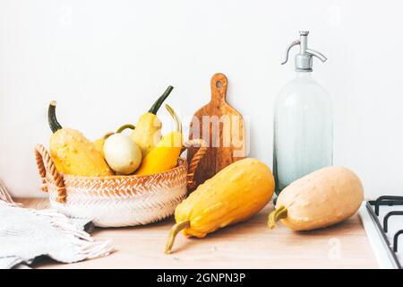 Küchentisch aus Holz mit bunten Kürbissen in Strickkorb, Carvingboard und Wasserflasche. Herbstliches Orange, gelbe Farben, Decke und Gasherd Stockfoto