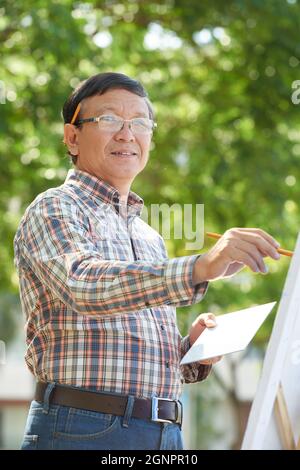 Porträt eines lächelnden reifen vietnamesischen Mannes in einer Brille, die im Park steht und ein Bild mit Farben zeichnet Stockfoto