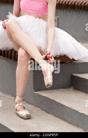 Die nicht erkennbare Ballettänzerin sitzt auf der Bank und zieht beige spitzenschuhe an. Sie trägt leichte Tanzbekleidung. Ballerina in der City Street Stockfoto