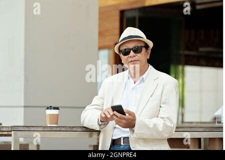 Porträt eines ernsthaften asiatischen Geschäftsmannes in Sonnenbrille und Strohhut, der mit einer Kaffeetasse im Café im Freien und mit einem Smartphone am Tisch steht Stockfoto