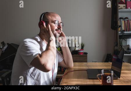 Junger Mann, der zu Hause mit einem Laptop arbeitet, eine Pause vom Musikhören macht. Mann hört gerne zu Hause Musik. Stockfoto