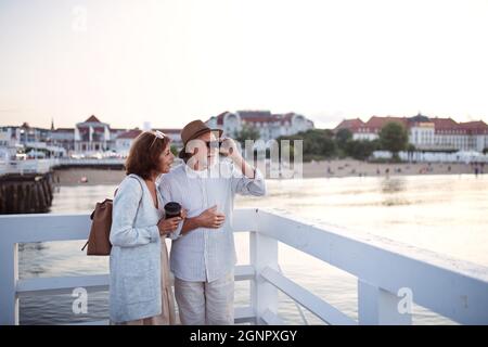 Ein glückliches Ehepaar umarmt sich draußen am Pier am Meer und blickt durch ein Fernglas auf die Aussicht. Stockfoto