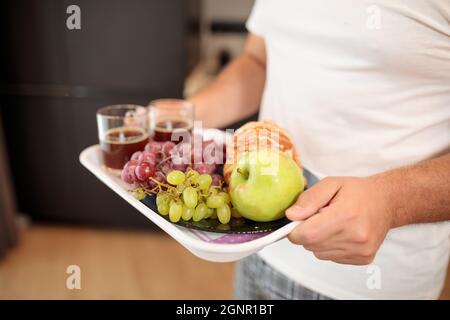 Gesunder Frühstückskaffee, Croissants, Graipes und Apfel. Männliche Hände halten einen Teller auf dem Hintergrund der Hausmannskost Stockfoto
