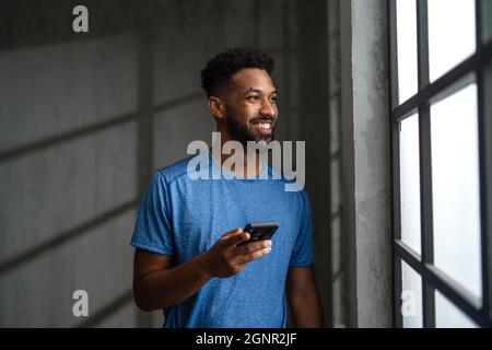 Glücklicher junger afroamerikanischer Sportler, der im Fitnessstudio mit dem Smartphone steht. Stockfoto