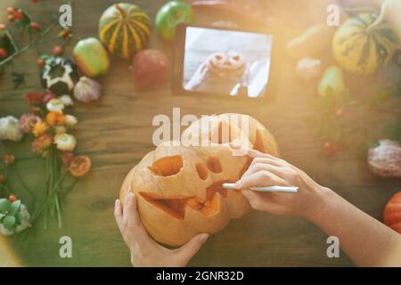 Halloween-Konzept: Frau schnitzt einen Mund in einen Kürbis, um eine Jack o'Laterne auf einem Holztisch mit Gemüse mit Video-Handbuch auf Tablet zu machen. Draufsicht. Hochwertige Bilder Stockfoto