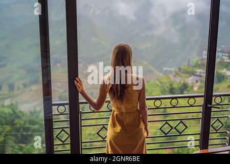 Eine junge Frau hält eine Kaffeetasse, während sie auf einem Stuhl auf dem Balkon sitzt und die Berge und die grüne Natur betrachtet. Frau in Sapa im Nebel, Nordwesten Stockfoto