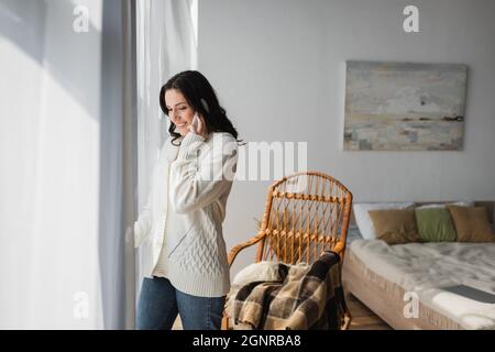 Lächelnde Frau, die auf dem Smartphone spricht, während sie in der Nähe des Fensters im Schlafzimmer steht Stockfoto