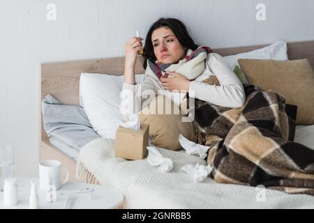 Aufgeregte Frau, die Nasenspray ansah, während sie im Bett in der Nähe von zerknitterten Papierservietten lag Stockfoto