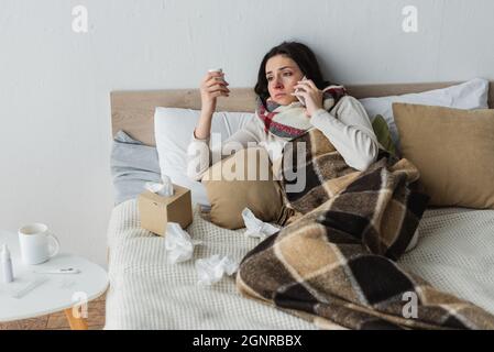 Kranke Frau mit Pillen Behälter reden auf Handy, während sie in der Nähe zerknittert Papier Servietten liegen Stockfoto