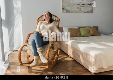 Fröhliche Frau in weichem Pullover und warmen Socken, die im Schaukelstuhl im Schlafzimmer ruhen Stockfoto