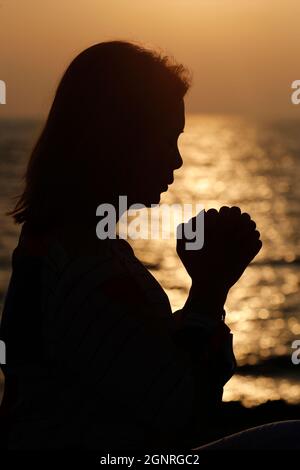 Silhouette bei Sonnenuntergang einer Gläubigen Frau, die betet, die Hände in Anbetung gottes gefaltet. Konzept für Religion, Glauben, Gebet und Spiritualität. Stockfoto