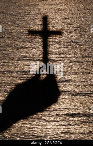 Gläubige Frau, die bei Sonnenuntergang mit Christenkreuz betet. Nahaufnahme auf einer Hand. Konzept für Religion, Glauben, Gebet und Spiritualität. Stockfoto