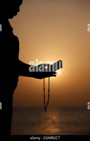 Silhouette einer treuen Frau, die bei Sonnenuntergang mit Rosenkranz betet. Konzept für Religion, Glauben, Gebet und Spiritualität. Stockfoto