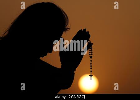 Silhouette einer treuen Frau, die bei Sonnenuntergang mit Rosenkranz betet. Konzept für Religion, Glauben, Gebet und Spiritualität. Stockfoto