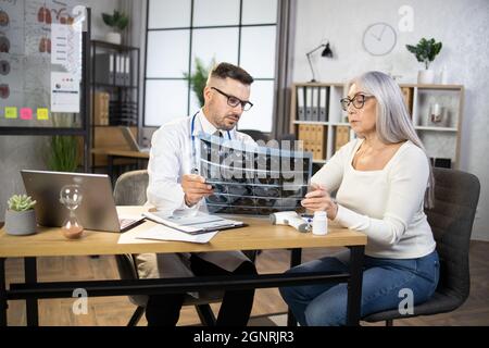 Konzentrierte ältere Frau, die mit einem professionellen kaukasischen Arzt am Schreibtisch sitzt und der Erklärung der Röntgentomographie zuhört. Weibliche Patientin, die im Krankenhaus ärztliche Beratung und Diagnose erhält. Stockfoto