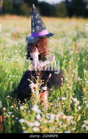 Ein Mädchen in einem schwarzen Kleid und einem Hexenhut sammelt Kräuter für einen Trank. Stockfoto