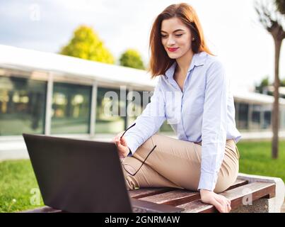 Junge Rotschopf kaukasische Frau lächelt in Laptop im Freien im Stadtpark Stockfoto