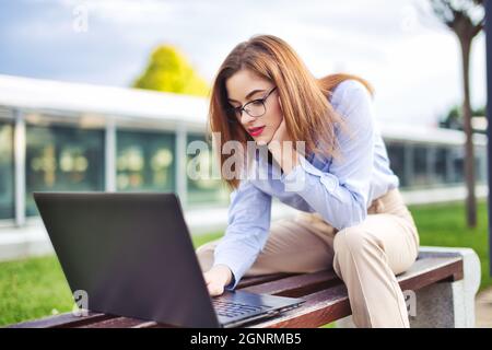 Junge Nerd Rotschopf kaukasische Frau mit Laptop im Freien, sitzt auf der Bank im Stadtpark Stockfoto