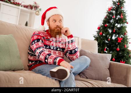 Foto von gut aussehenden ernst männlichen Look seitwärts denken Brainstorming tragen santa Hut hässlich Pullover Pullover sitzen Sofa zu Hause Stockfoto