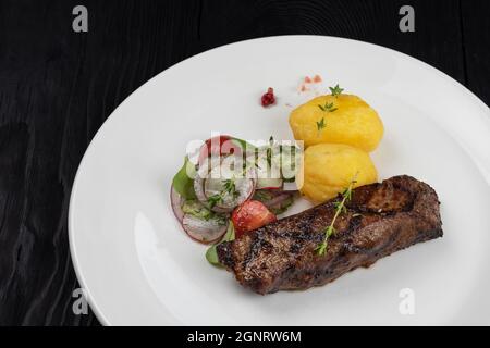 Gegrilltes Rindsrock-Steak mit Kartoffeln und Gemüse auf Weiße Platte auf schwarzem Holzhintergrund Stockfoto