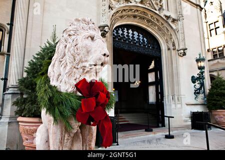 Eine mit einem Weihnachtsbogen geschmückte Statue eines Schutzlöwens am Eingang des Biltmore Estate, das im Herbst in Asheville, North Carolina, im Privatbesitz der Familie Vanderbilt ist. Das Haus ist das größte Privathaus in Amerika mit über 250 Zimmern. Stockfoto