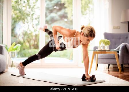 Aufnahme einer Frau, die Hanteln verwendet, während sie zu Hause ein Körpertraining macht. Frau, die Sportkleidung trägt, während sie auf der Yogamatte trainiert. Gesunder Lebensstil. Stockfoto