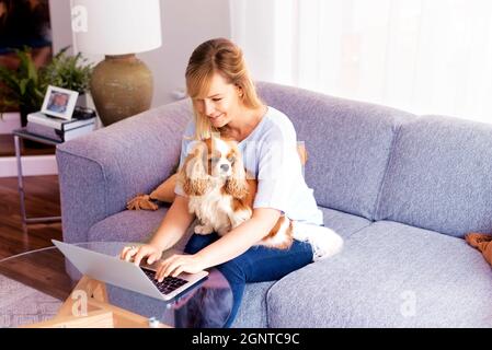Aufnahme einer Geschäftsfrau mittleren Alters, die ihren Laptop benutzt, während sie zu Hause auf einer Couch sitzt und von zu Hause aus arbeitet. Ihr süßer Welpe entspannt sich in ihrem Schoß. Stockfoto