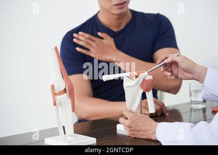 Horizontale Aufnahme eines Traumatologen, der vor einem jungen asiatischen Mann am Schreibtisch sitzt und ihn wegen Schmerzen im Schultergelenk konsultiert Stockfoto