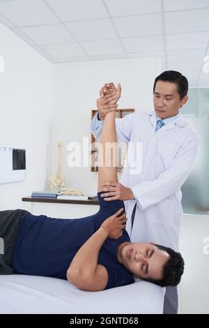 Vertikale Aufnahme eines reifen asiatischen Physiotherapeuten, der mit einem jungen männlichen Patienten arbeitet, der nach einer Verletzung seine Flexibilität am Schultergelenk wiedererlangt Stockfoto