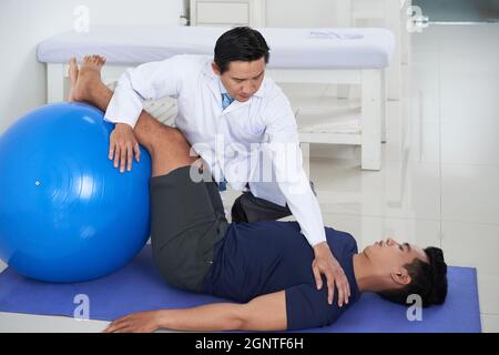 Horizontale Aufnahme eines modernen asiatischen Physiotherapeuten, der männlichen Patienten bei der Ausübung von Übungen auf der Matte mit Balleinsatz zur Rehabilitation unterstützt Stockfoto
