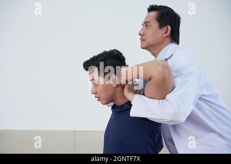 Horizontale mittlere Seitenansicht Aufnahme eines modernen reiferen asiatischen Osteopathietherapeuten, der junge Männer behandelt, die seine Knochen und Muskeln manipulieren Stockfoto