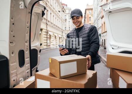Lächelnder Kurier in Uniform, der am Kofferraum des Autos stand und das Mobiltelefon auf Kartons blickte Stockfoto