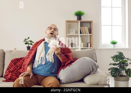 Kranker Mann, der zu Hause mit Serviette in den Händen sitzt, niest wegen Erkältung oder Grippevirus. Stockfoto