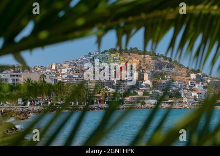 Kusadasi, Aydin, Türkei - 22. August 2021: Farbenfrohe Kusadasi-Häuser auf dem Hügel, durch Palmblätter gesehen Stockfoto