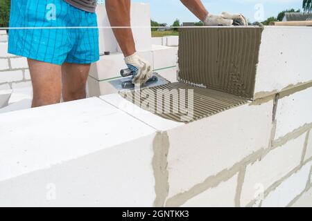 Der Baumeister verwendet eine gekerbte Kelle, um das Zementgemisch aus nächster Nähe auf die belüftelten Betonblöcke aufzutragen. Konzept des Aufbaus eines Privathauses Stockfoto