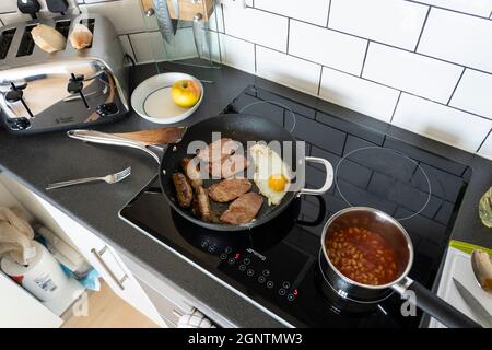 Traditionelles englisches Frühstück in einer Küche mit einem Spiegelei, Speckscheiben und Würstchen, die in einer Bratpfanne zubereitet werden, und gebackenen Bohnen in einem Topf. England Stockfoto