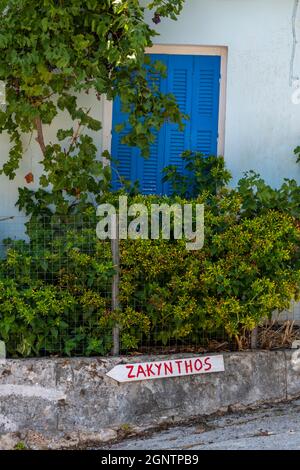 Blau lackierte Holztür auf einem weiß gestrichenen griechischen Gebäude mit handgemaltem Straßenschild für die Stadt zakynthos, das an der Außenwand der Straße befestigt ist Stockfoto