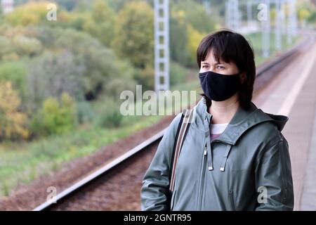 Frau in schwarzer Maske wartet auf einen Zug auf einem Bahnhof. Sicherheitsmaßnahmen für den öffentlichen Verkehr während der Pandemie Covid-19 Stockfoto