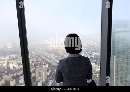 Frau, die vor einem Panoramafenster eines Wolkenkratzers steht und auf die neblige Stadt hinunterblickt Stockfoto