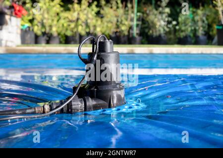 Automatische Pumpe für die Poolabdeckung auf der blauen Nassabdeckung vor unbelaubten Gartenbeläumungen. Perspektivische Ansicht der Pumpe der schwarzen Poolabdeckung zum Halten des Wattes Stockfoto