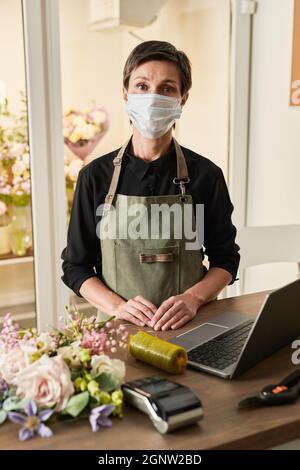 Vertikales Porträt einer weiblichen Verkäuferin, die eine Maske trägt, während sie am Kassierstisch im Blumenladen steht Stockfoto