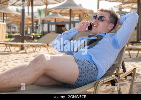 Der junge glückliche Geschäftsmann downshifter spricht am Telefon und liegt auf einer Sonnenliege mitten am Strand in einem Hemd, Krawatte und Boxershorts Stockfoto