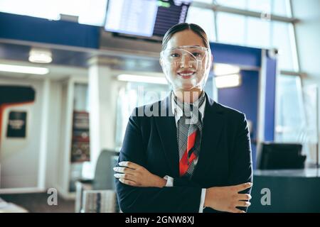 Porträt einer weiblichen Bodenbegleiterin, die während einer Pandemie arbeitet. Frau mit Gesichtsschutz, die mit gekreuzten Armen am Flughafenterminal steht. Stockfoto