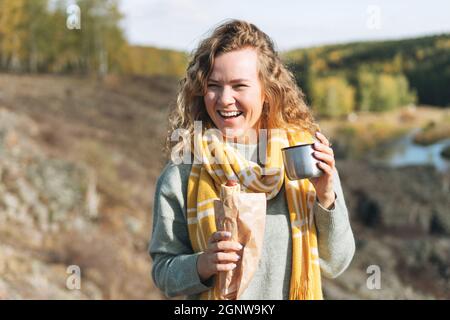 Junge glückliche schöne Frau Reisende mit lockigen Haaren essen Hot Dog und trinken Tee auf dem Hintergrund der Berge und Fluss, Wandern im Herbst natu Stockfoto