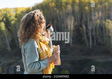 Junge glückliche schöne Frau Reisende mit lockigen Haaren essen Hot Dog und trinken Tee auf dem Hintergrund der Berge und Fluss, Wandern im Herbst natu Stockfoto