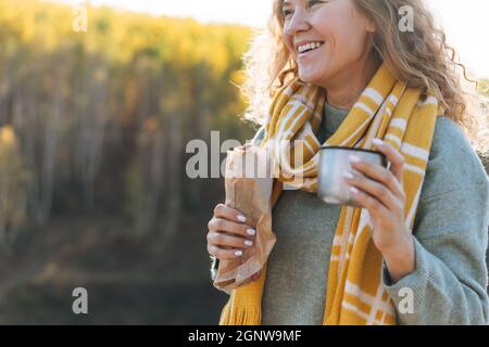 Junge glückliche schöne Frau Reisende mit lockigen Haaren essen Hot Dog und trinken Tee auf dem Hintergrund der Berge und Fluss, Wandern im Herbst natu Stockfoto