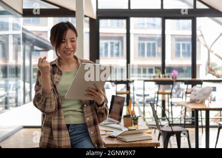 Junge asiatische Frau feiern Erfolg oder glückliche Pose während der Hand halten digitale Tablet, Frau in ihrem Büro arbeiten Stockfoto