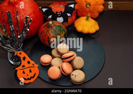 Dessert für Halloween. Erntedankfest-Menü. Französische Macarons. Makronen mit Schokolade auf schwarzem Hintergrund mit Kürbissen und Öko-Cocktail-Trinkhalmen. Weich Stockfoto