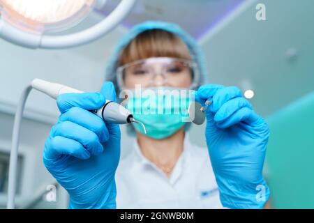 Zahnarzt in einer OP-Maske, hält einen Winkelspiegel und einen Bohrer, bereit zur Arbeit. Frau Zahnärztin bei der Arbeit. Macht eine Untersuchung und Behandlung von Zähnen für Karies. Stockfoto